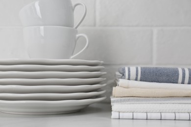 Stack of soft kitchen towels and dishware on table near white brick wall, closeup