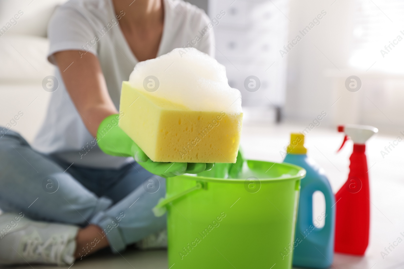 Photo of Woman holding sponge with foam over bucket indoors, closeup. Cleaning supplies