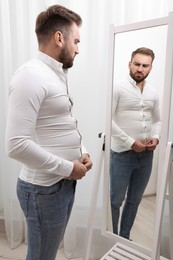 Photo of Upset man wearing tight shirt in front of mirror at home. Overweight problem