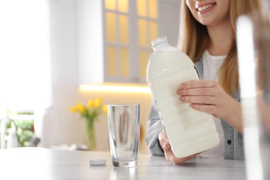 Young woman pouring milk from gallon bottle into glass at white marble table in kitchen, closeup