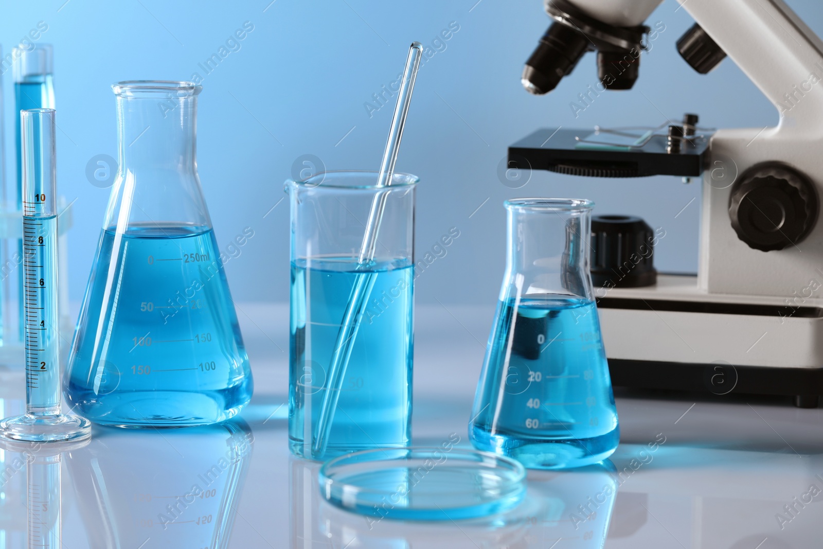 Photo of Different laboratory glassware with light blue liquid and microscope on table