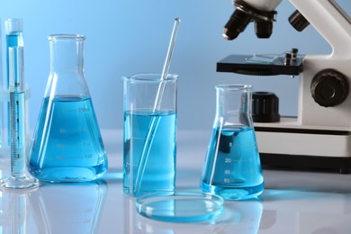 Photo of Different laboratory glassware with light blue liquid and microscope on table