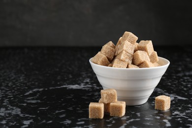 Photo of Brown sugar cubes in bowl on dark textured table, closeup. Space for text