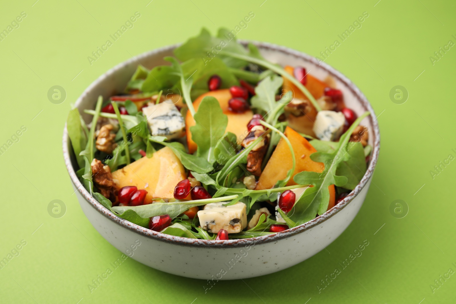 Photo of Tasty salad with persimmon, blue cheese, pomegranate and walnuts served on light green background