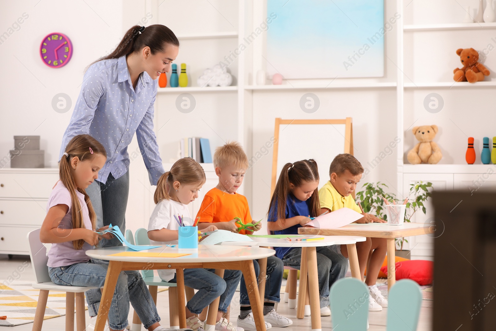 Photo of Nursery teacher with group of cute little children drawing and cutting paper at desks in kindergarten. Playtime activities