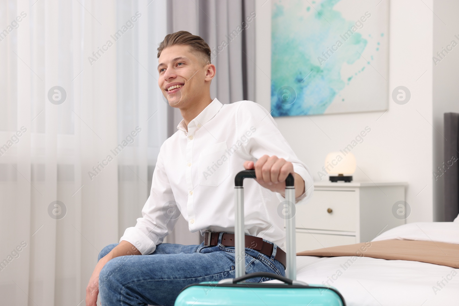 Photo of Smiling guest with suitcase relaxing on bed in stylish hotel room