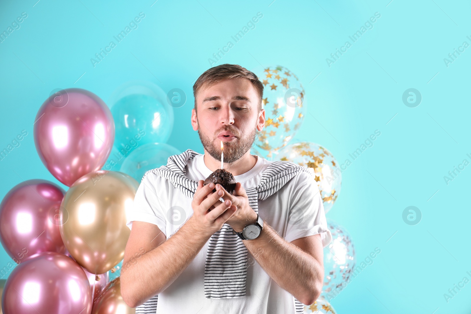 Photo of Young man with birthday muffin and air balloons on color background