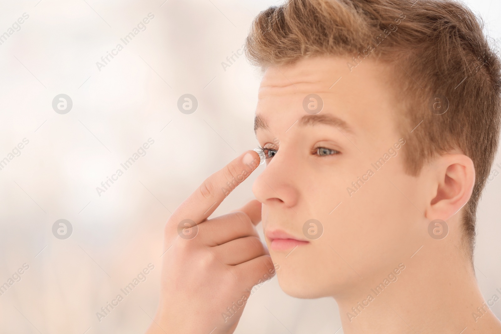 Photo of Teenage boy putting contact lens in his eye on blurred background