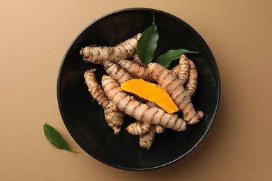 Bowl with raw turmeric roots and green leaves on light brown table, flat lay