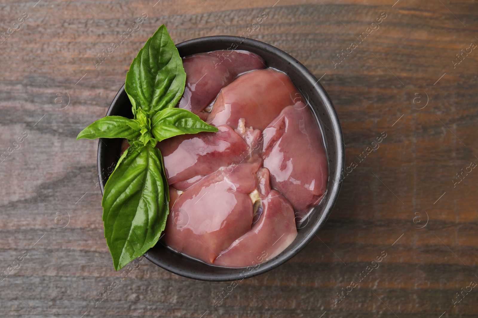 Photo of Bowl of raw chicken liver with basil on wooden table, top view