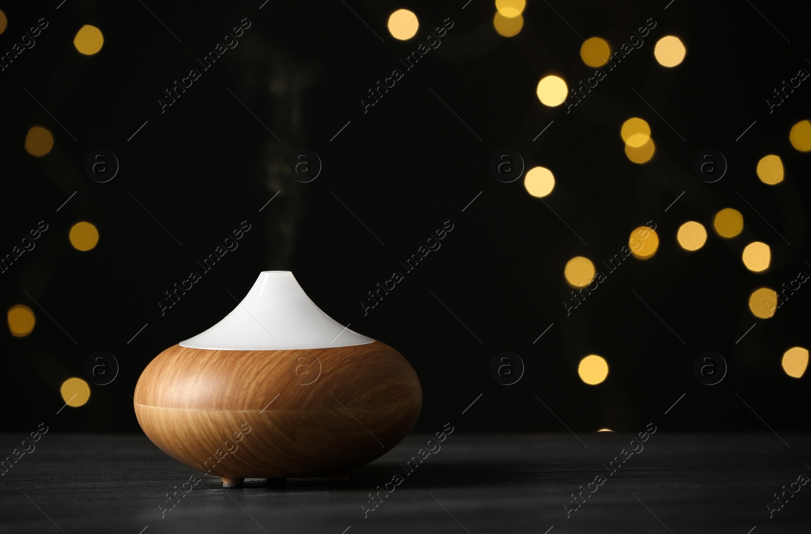 Photo of Modern essential oil diffuser on wooden table against blurred lights, space for text