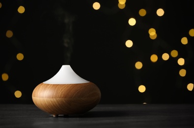 Modern essential oil diffuser on wooden table against blurred lights, space for text