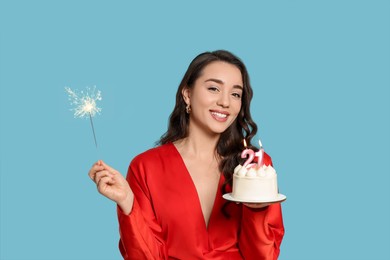 Photo of Coming of age party - 21st birthday. Woman holding sparkler and delicious cake with number shaped candles on light blue background