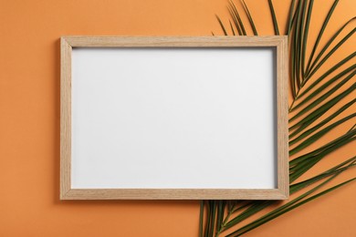 Photo of Blank photo frame and tropical leaf on orange background, flat lay