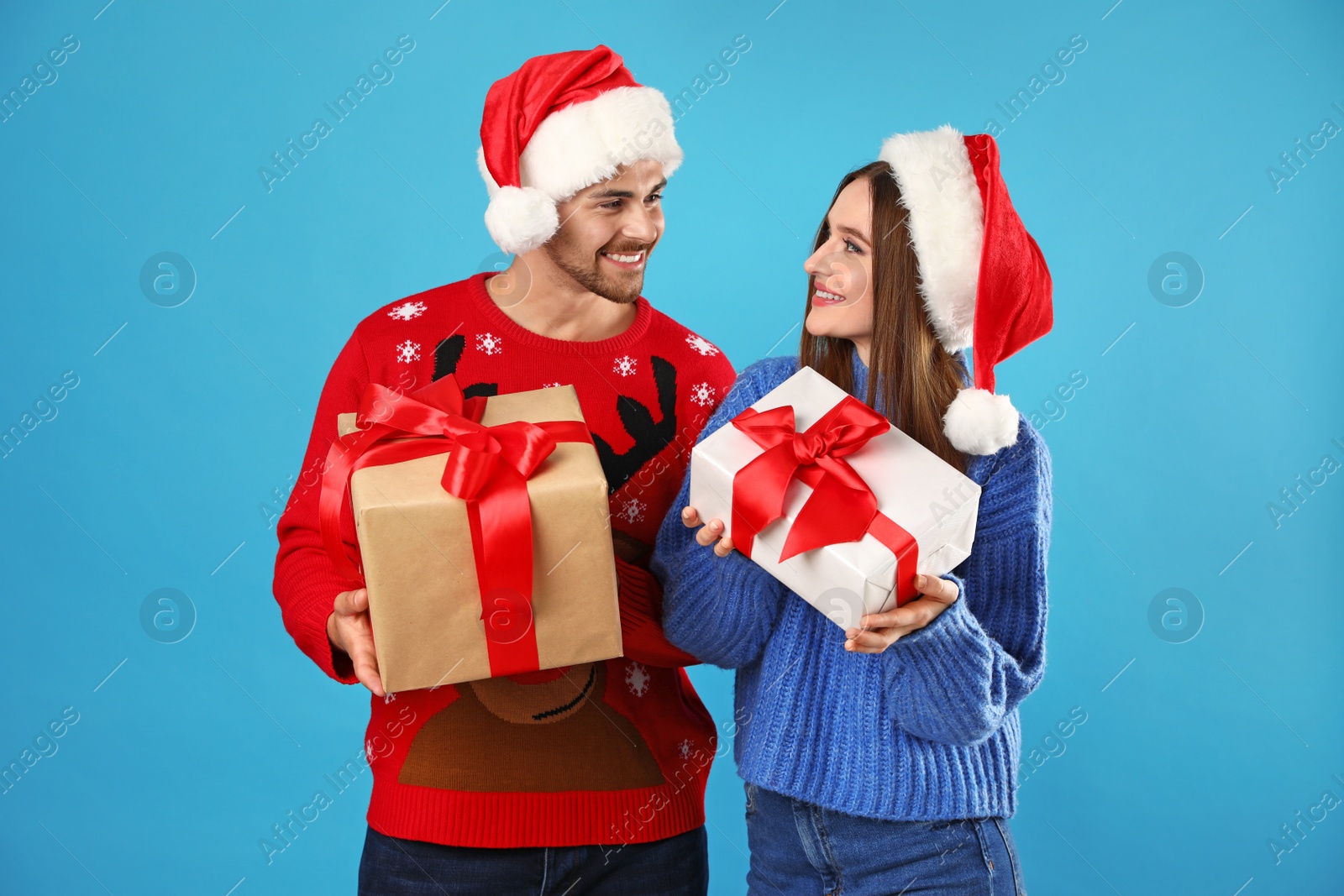 Photo of Couple in Christmas sweaters with Santa hats and gift boxes on blue background