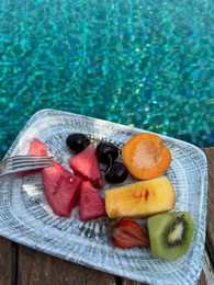 Photo of Plate with fresh fruits on wooden deck near outdoor swimming pool. Luxury resort