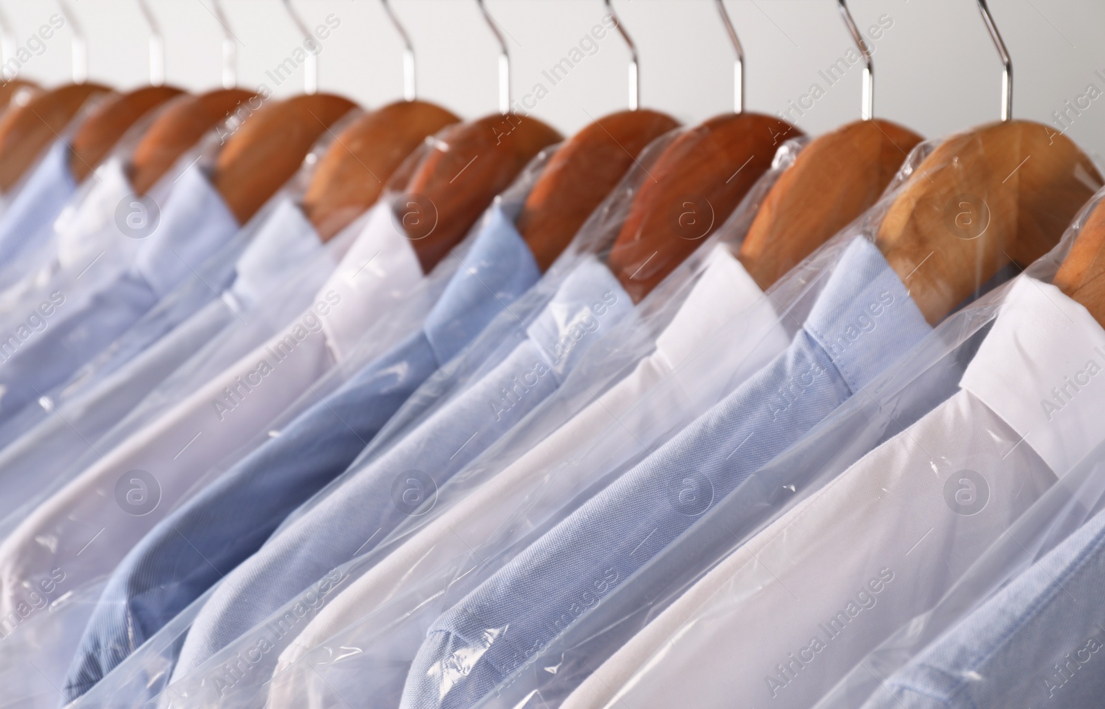 Photo of Hangers with shirts in dry cleaning plastic bags on rack against light background, closeup