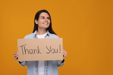 Photo of Happy woman holding cardboard sheet with phrase Thank You on orange background, space for text