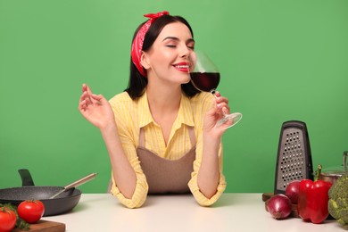 Photo of Young housewife with glass of wine, vegetables and different utensils on green background