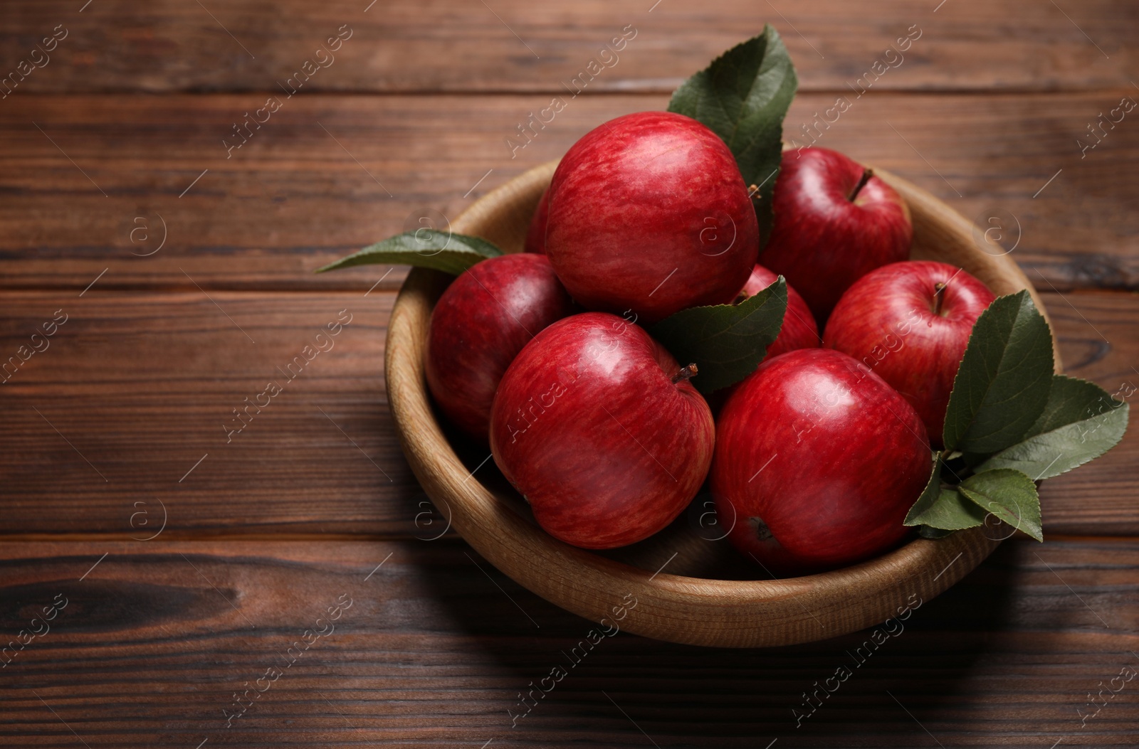 Photo of Ripe red apples and green leaves in bowl on wooden table. Space for text