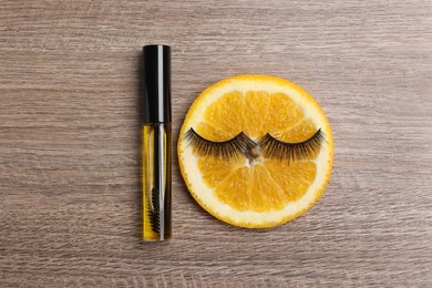 False eyelashes, orange slice and tube of oil on wooden table, flat lay