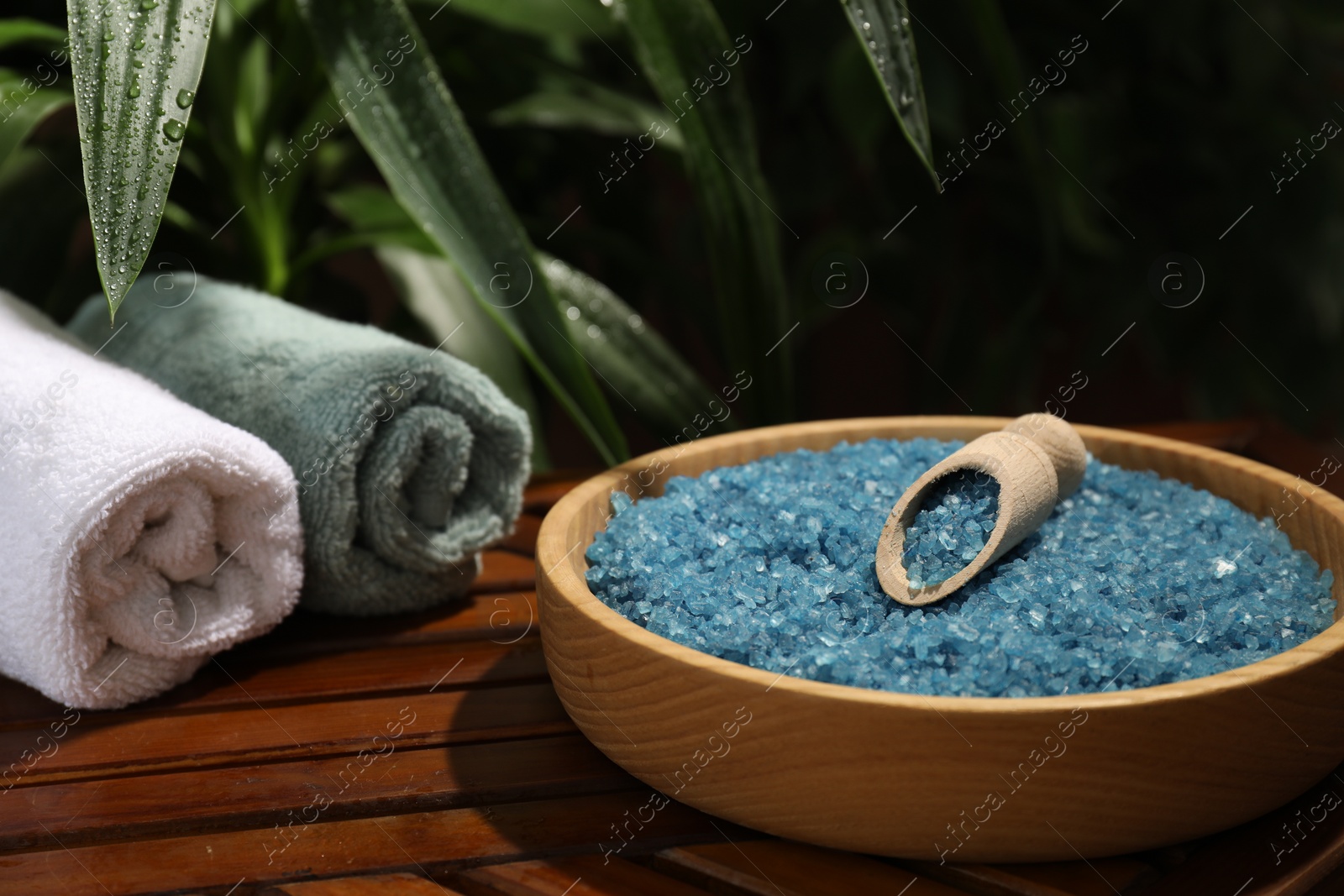 Photo of Bowl of blue sea salt with scoop and rolled towels on wooden table, closeup