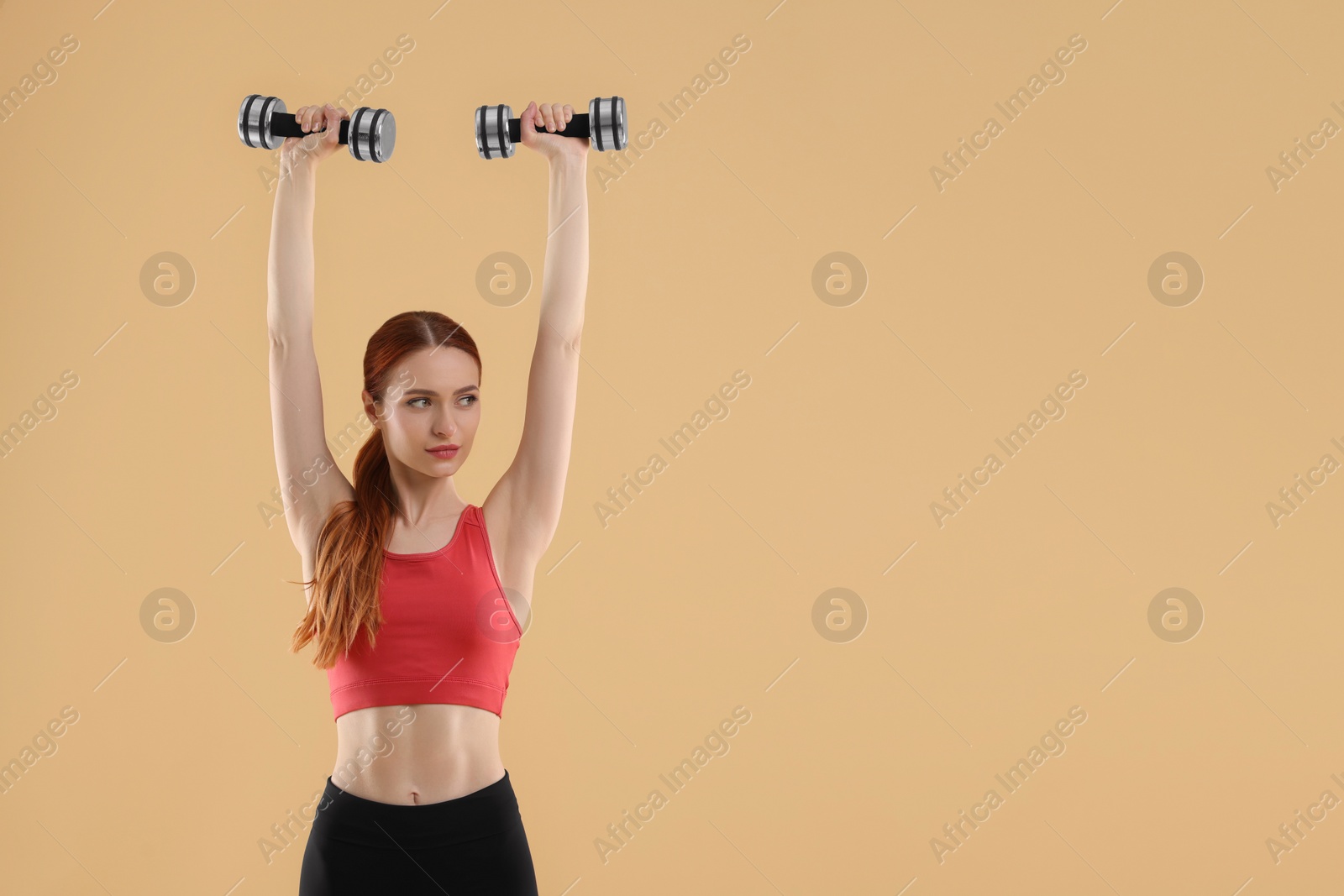 Photo of Young woman in sportswear doing exercises with dumbbells on beige background, space for text