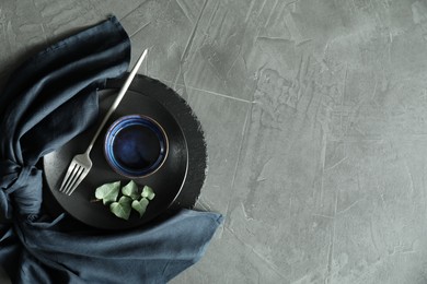 Stylish table setting. Dishes, fork, napkin and green leaves on grey surface, top view with space for text