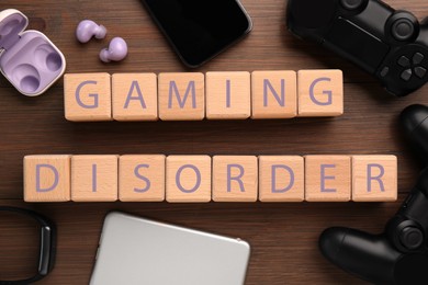 Photo of Phrase Gaming Disorder made of cubes and different devices on wooden table, flat lay. Addictive behavior