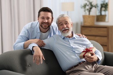 Photo of Son giving gift box to his dad on sofa at home