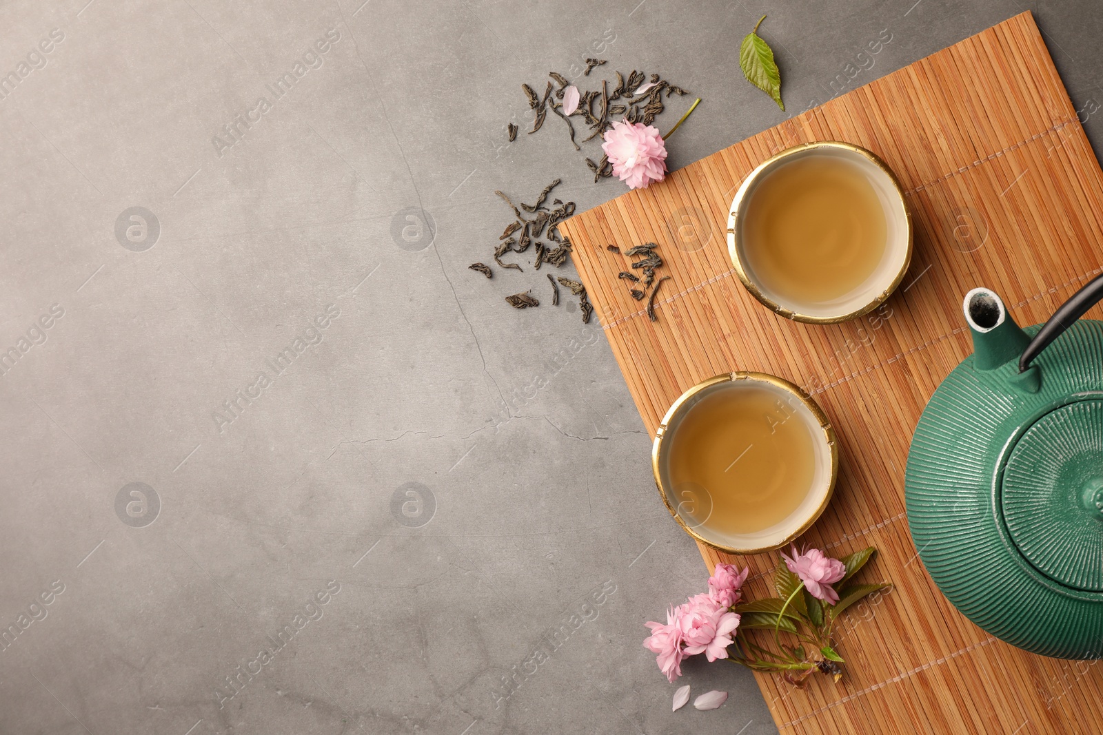 Photo of Traditional ceremony. Cups of brewed tea, teapot and sakura flowers on grey table, flat lay with space for text