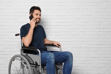 Young man in wheelchair talking on phone near brick wall indoors. Space for text