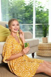 Photo of Young woman with glass of lemonade talking on phone at home. Refreshing drink