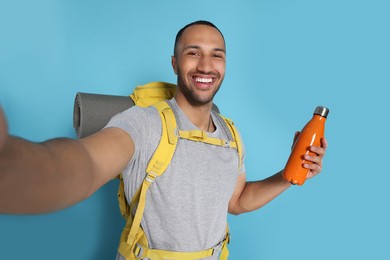 Photo of Happy tourist with backpack and thermo bottle taking selfie on light blue background