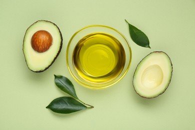 Photo of Bowl with oil, fresh cut avocados and leaves on light green background, flat lay