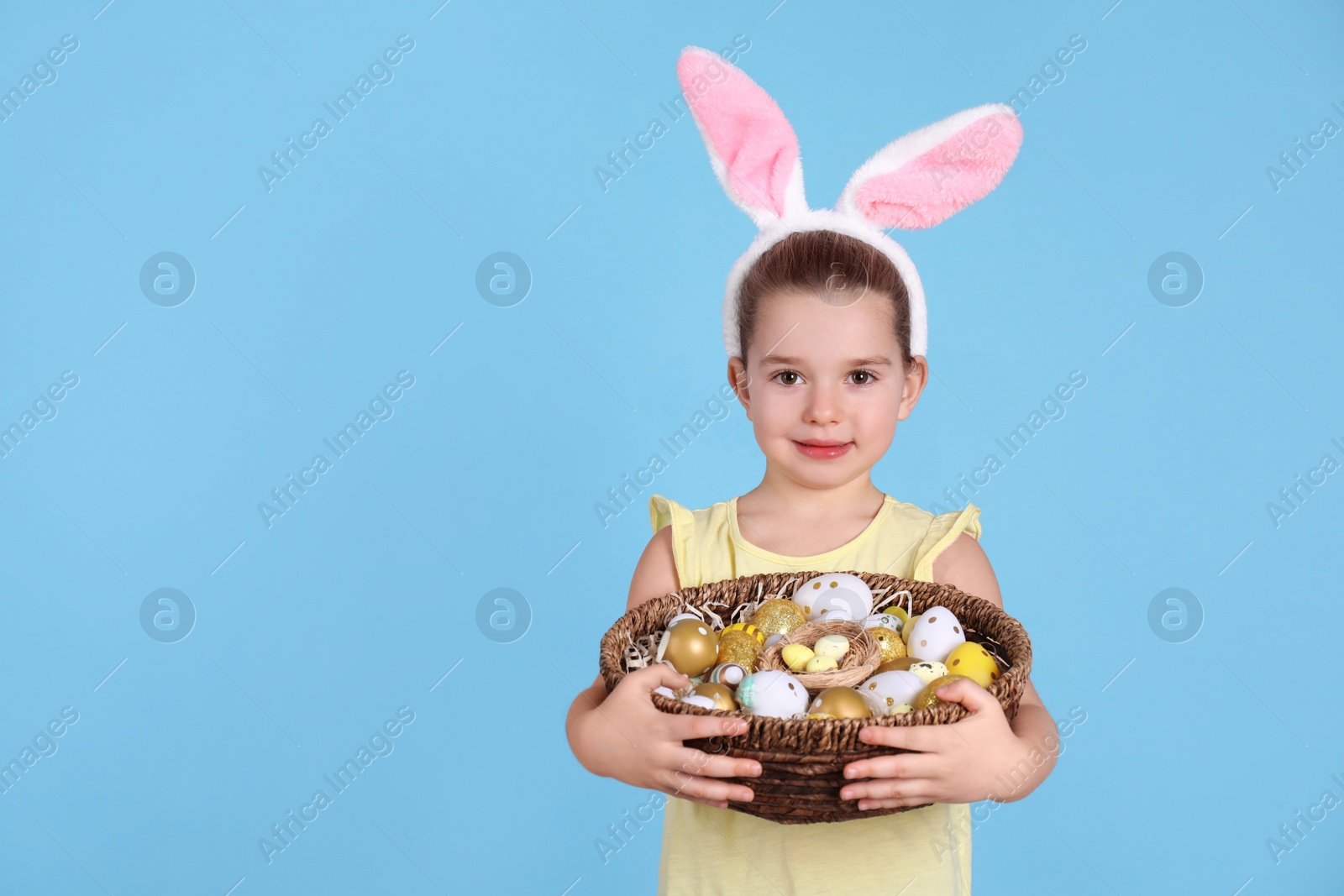 Photo of Happy little girl with bunny ears holding wicker basket full of Easter eggs on light blue background. Space for text