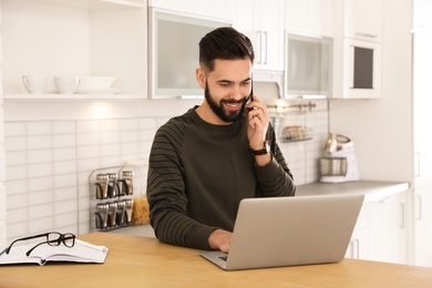 Handsome young man talking on phone while working at table in kitchen