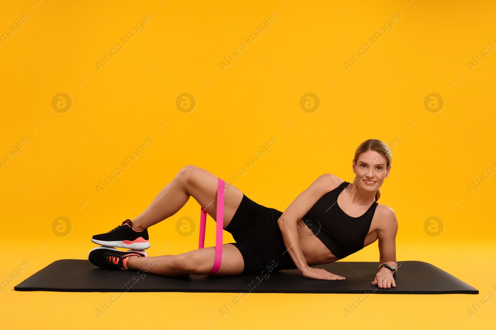 Photo of Woman exercising with elastic resistance band on fitness mat against orange background