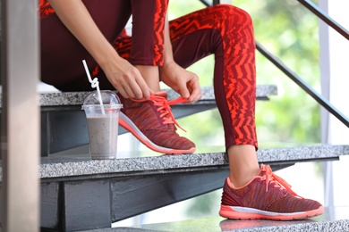 Young woman in sportswear with plastic cup of healthy smoothie on stairs indoors