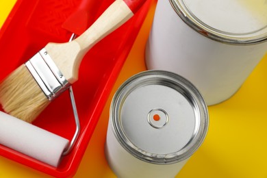 Photo of Cans of orange paint, brush, roller and container on yellow background, closeup