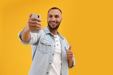 Photo of Smiling young man taking selfie with smartphone and showing thumbs up on yellow background, space for text