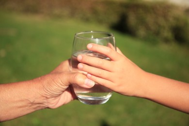 Photo of Child giving glass of water to elderly woman outdoors on sunny day, closeup