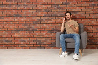 Man talking on smartphone in armchair near brick wall, space for text. Interior design