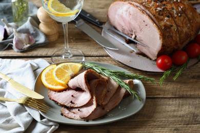Photo of Slices of delicious baked ham, orange, rosemary, fork, and knife on wooden table