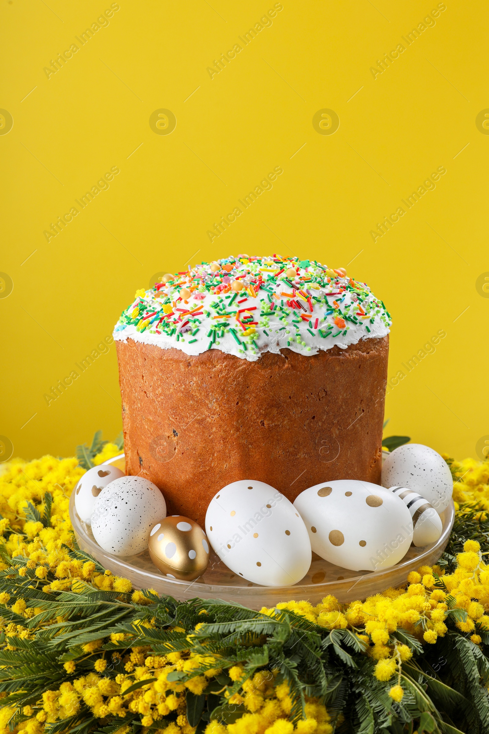 Photo of Traditional Easter cake with sprinkles, painted eggs and beautiful spring flowers on yellow background, closeup