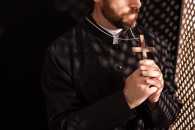 Catholic priest in cassock holding cross in confessional booth, closeup