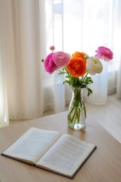 Photo of Bouquet of beautiful ranunculus flowers in vase and open book on wooden table indoors