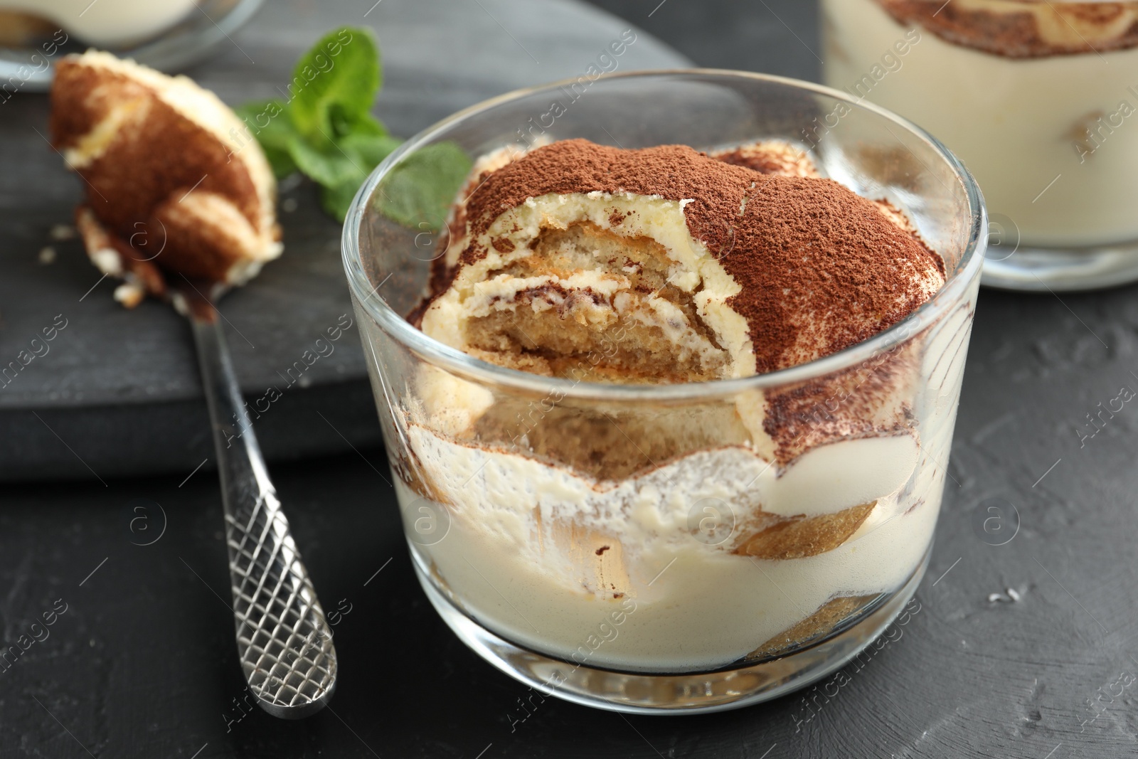 Photo of Delicious tiramisu in glass and spoon on black table, closeup