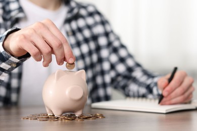 Photo of Financial savings. Man putting coin into piggy bank while writing down notes at wooden table, closeup