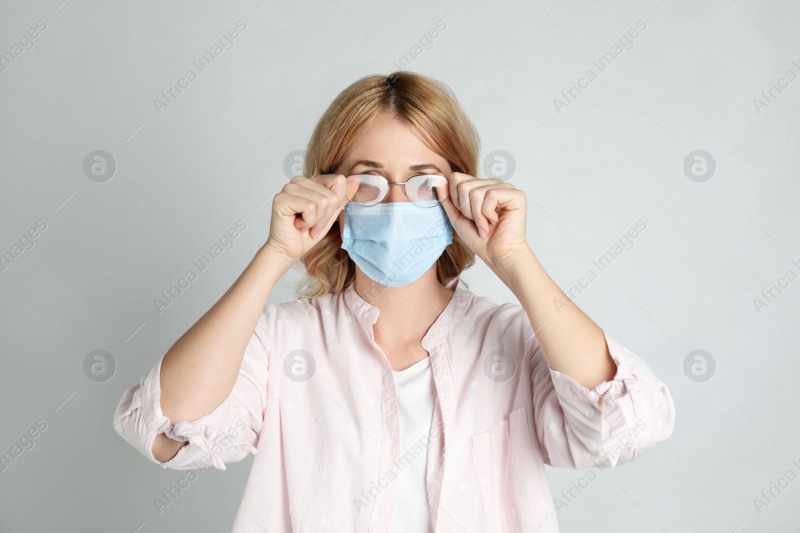 Photo of Woman wiping foggy glasses caused by wearing medical mask on light background